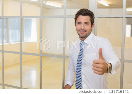 Man standing in office by glass panes giving thumbs-up wearing white shirt striped tie, copy space 135716141