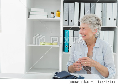 Senior female in button-up shirt smiling, holding hands, sitting at desk with binders, copy space Senior female in button-up shirt smiling, holding hands, sitting at desk with binders, copy space 135716143