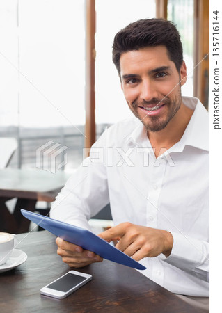 Man in his 30s sitting in cafe wearing button-up shirt holding blue tablet tapping, copy space Man in his 30s sitting in cafe wearing button-up shirt holding blue tablet tapping, copy space 135716144