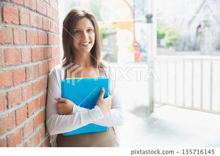 Teenage female student leaning at building entrance against red brick wall, holding blue binder 135716145
