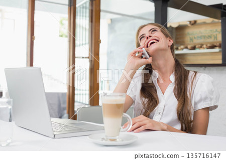 Woman sitting at cafe wearing shirt, talking on phone, laughing at laptop, latte, copy space Woman sitting at cafe wearing shirt, talking on phone, laughing at laptop, latte, copy space 135716147