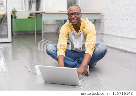 African American man sitting in studio working on silver laptop, yellow hoodie, glasses, coffee cup 135716148