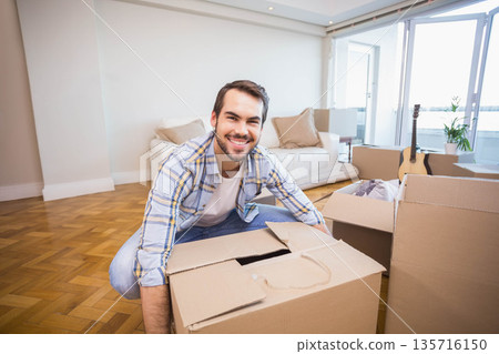 Male kneeling, unpacking moving boxes in living room wearing plaid shirt and jeans near guitar Male kneeling, unpacking moving boxes in living room wearing plaid shirt and jeans near guitar 135716150