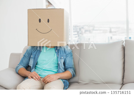 Adult male sitting on light-gray sofa in living room by window wearing denim shirt cardboard box 135716151