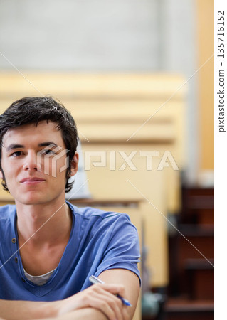 Male in twenties sitting at lecture hall desk wearing blue short-sleeve shirt and holding pen Male in twenties sitting at lecture hall desk wearing blue short-sleeve shirt and holding pen 135716152