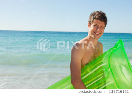 Mid-adult man standing in surf at sunny beach holding green inflatable float shirtless, copy space 135716161