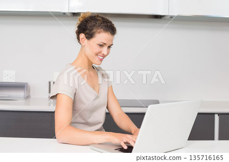 Adult woman wearing light beige T-shirt sitting at home kitchen countertop typing on silver laptop Adult woman wearing light beige T-shirt sitting at home kitchen countertop typing on silver laptop 135716165