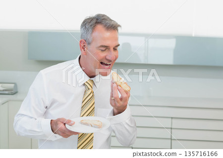 Mature man standing in kitchen wearing white shirt striped tie holding plate with sandwich 135716166