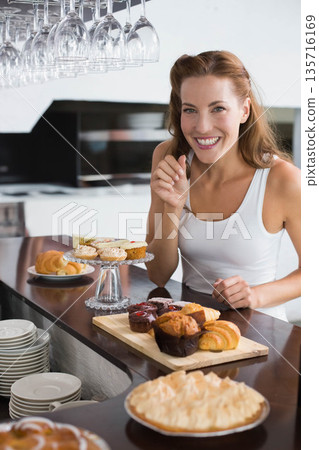 Mid-adult female standing behind polished wood counter, smiling gesturing near cupcakes, copy space 135716169
