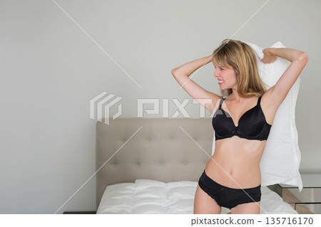 Woman in black bra and underwear standing on bed holding white pillow overhead bedroom, copy space 135716170