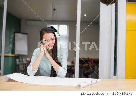 Asian woman in her twenties wearing light green shirt leaning over blueprint at modern workspace 135716175