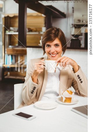 Adult female sitting at cafe table wearing blazer holding coffee cup and muffin smiling at camera Adult female sitting at cafe table wearing blazer holding coffee cup and muffin smiling at camera 135716177