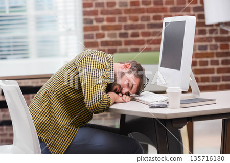 Adult African American man resting head on desk near monitor in office wearing checkered shirt Adult African American man resting head on desk near monitor in office wearing checkered shirt 135716180