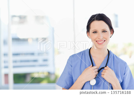 Adult female clinician standing, smiling, in blue scrubs holding stethoscope in clinic, copy space 135716188
