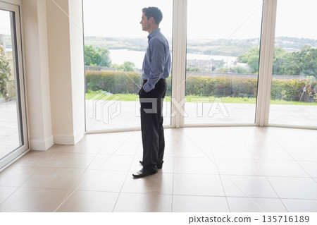 Businessman standing in modern living space, looking out floor-to-ceiling windows at waterfront 135716189