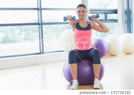 Adult female sitting on purple stability ball in gym holding blue dumbbells wearing pink tank top Adult female sitting on purple stability ball in gym holding blue dumbbells wearing pink tank top 135716192