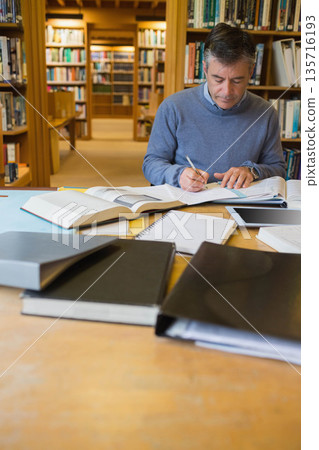 Man sitting at wooden study table in library, taking notes with open books, pen, tablet 135716193