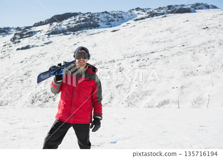 Adult man standing on alpine slope carrying skis on right shoulder, wearing goggles beanie gloves Adult man standing on alpine slope carrying skis on right shoulder, wearing goggles beanie gloves 135716194