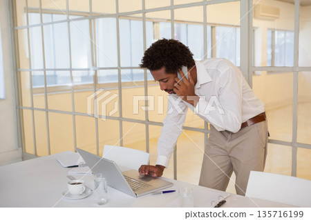 African American man in white shirt leaning over silver laptop holding phone in office, copy space African American man in white shirt leaning over silver laptop holding phone in office, copy space 135716199
