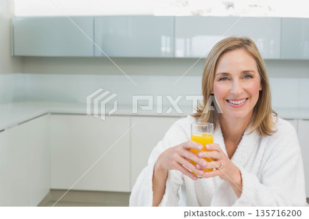 Mature woman smiling and holding glass of orange juice on countertop in bathrobe, copy space Mature woman smiling and holding glass of orange juice on countertop in bathrobe, copy space 135716200