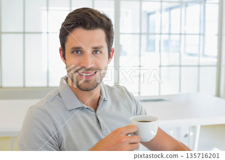 Man in late-20s sitting at white desk near grid-windows wearing gray polo holding white coffee cup 135716201