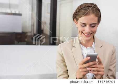 Woman sitting on bench in office wearing beige blazer and white top holding smartphone, copy space 135716204