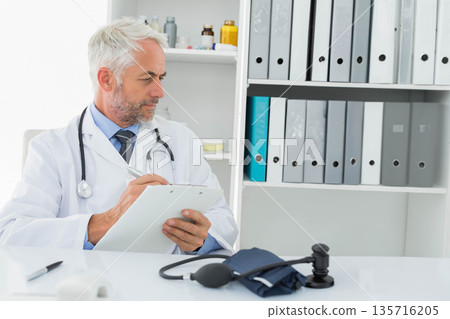 Senior male physician sitting at desk in clinic wearing lab coat, holding stethoscope and clipboard 135716205