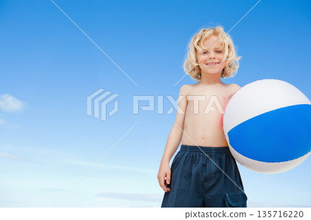 Boy child standing right on beach holding white blue beach ball in dark trunks, copy space 135716220