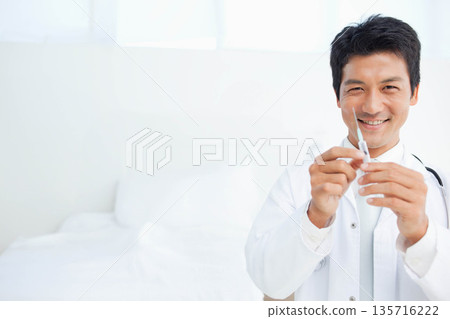Asian mid-adult man wearing white coat and stethoscope, holding syringe near exam bed, copy space 135716222