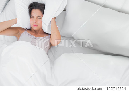 Woman lying on white bedding wearing camisole, holding pillow over ears, blocking noise, copy space Woman lying on white bedding wearing camisole, holding pillow over ears, blocking noise, copy space 135716224