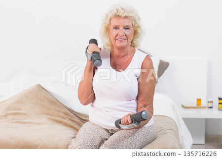 Senior woman sitting on bed in white tank top lifting small dumbbells near nightstand meds 135716226
