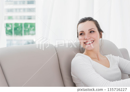 20s woman wearing white top sitting on light-gray sofa by large window, sheer curtains, copy space 135716227