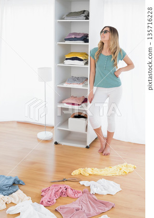 Woman standing barefoot by shelf wearing sunglasses, teal top and white cropped pants, copy space 135716228