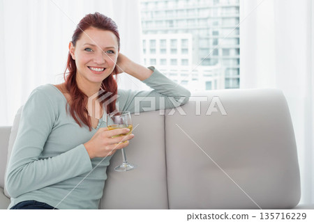 Woman sitting in living room wearing light green top holding stemmed wine glass, smiling by window 135716229