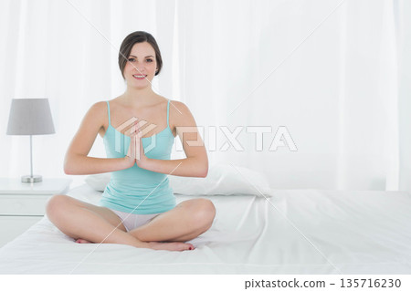 Woman sitting on bed praying wearing teal top with bedside lamp and sheer curtains filtering light 135716230