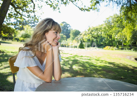 Woman sitting at round outdoor table on chair in park, wearing white short-sleeve dress, smiling Woman sitting at round outdoor table on chair in park, wearing white short-sleeve dress, smiling 135716234