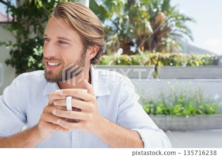 Man sitting on sunny terrace holding white ceramic mug at table, wearing shirt, planters with palms 135716238