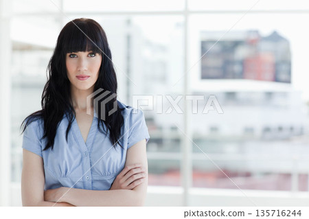 Adult woman standing facing camera with arms crossed in light blue blouse at office windows 135716244