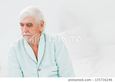 Senior man sitting on bed in bright bedroom wearing seafoam blue robe, touching white pillows Senior man sitting on bed in bright bedroom wearing seafoam blue robe, touching white pillows 135716246