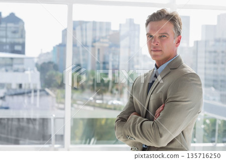 Mature man standing arms crossed by glass window showing skyline, in suit and tie, copy space 135716250