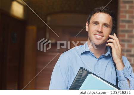 Mid-30s man talking on phone in hall, light-blue shirt, portfolio with blue paper, copy space 135716251
