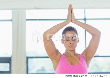 Female doing yoga with arms raised, wearing pink athletic tank top in bright studio with windows 135716256