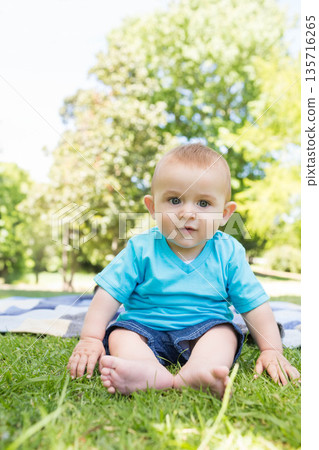 Infant boy sitting on striped picnic blanket in park wearing turquoise shirt and denim shorts Infant boy sitting on striped picnic blanket in park wearing turquoise shirt and denim shorts 135716265