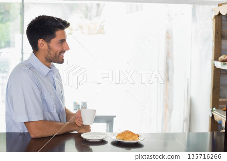 Man sitting at counter in cafe wearing light blue shirt holding white mug with croissant, smiling Man sitting at counter in cafe wearing light blue shirt holding white mug with croissant, smiling 135716266