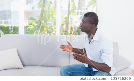 Adult African American man in light shirt, jeans sitting on sofa at home gesturing, copy space Adult African American man in light shirt, jeans sitting on sofa at home gesturing, copy space 135716269