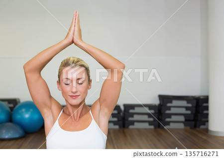 Woman doing yoga in studio wearing white tank top, standing by stacked steps and blue balls Woman doing yoga in studio wearing white tank top, standing by stacked steps and blue balls 135716271