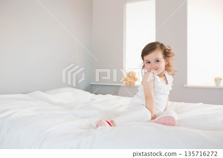 Female toddler sitting on white bed in bedroom with teddy bear wearing white top, copy space Female toddler sitting on white bed in bedroom with teddy bear wearing white top, copy space 135716272