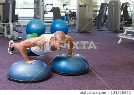 Male in late 20s doing push-ups on blue BOSU-style trainers at gym wearing white tank top 135716273