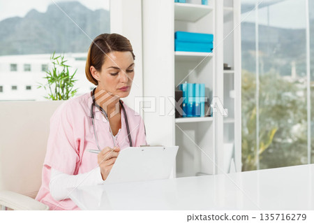 Female in pink scrubs sitting at desk in medical office writing on clipboard wearing stethoscope 135716279