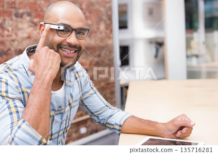 Mid adult man in plaid shirt tapping AR-display on glasses, sitting at loft table with tablet Mid adult man in plaid shirt tapping AR-display on glasses, sitting at loft table with tablet 135716281